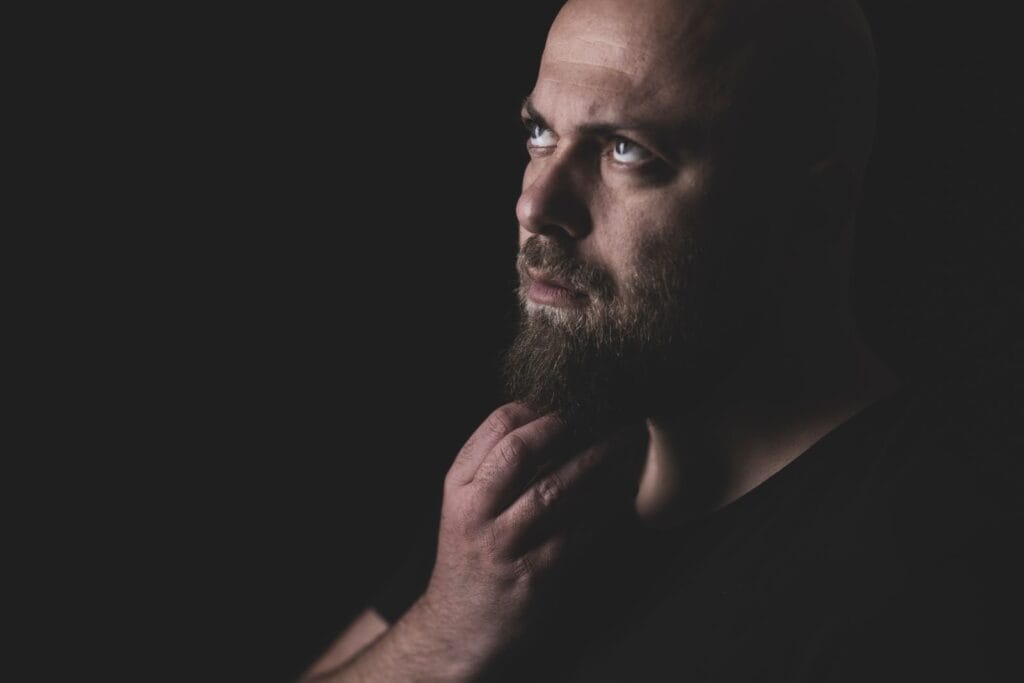 A close-up portrait of a bald man with a beard, looking contemplatively upwards in a studio setting.
