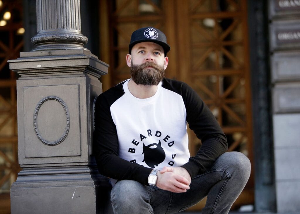 Stylish bearded man in cap and casual wear sitting in city street scene.