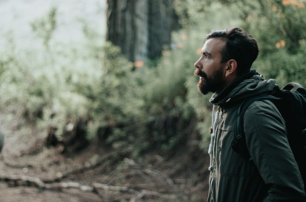A bearded man in a green jacket hiking and exploring a forest.