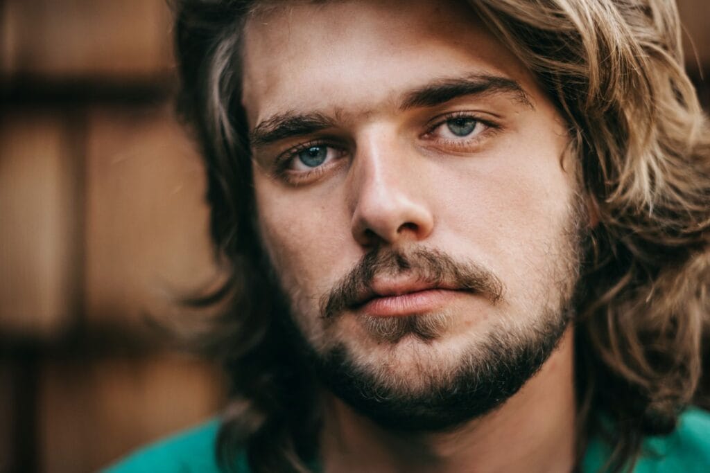 A striking close-up portrait of a young bearded man with long hair and blue eyes.