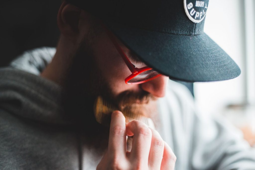High angle of modern man in stylish cap and glasses combing beard while resting at home